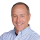 Professional headshot of a smiling man wearing a light blue striped button-up shirt against a white background